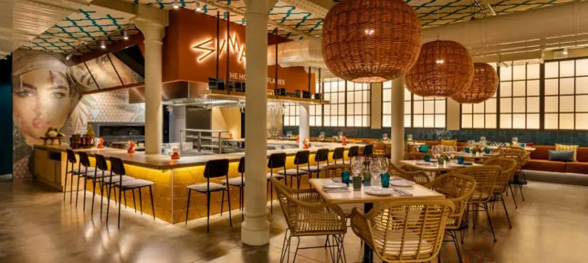 Restaurante con barra iluminada, grandes lámparas de fibra natural, columnas de fundición y sillas de rejilla