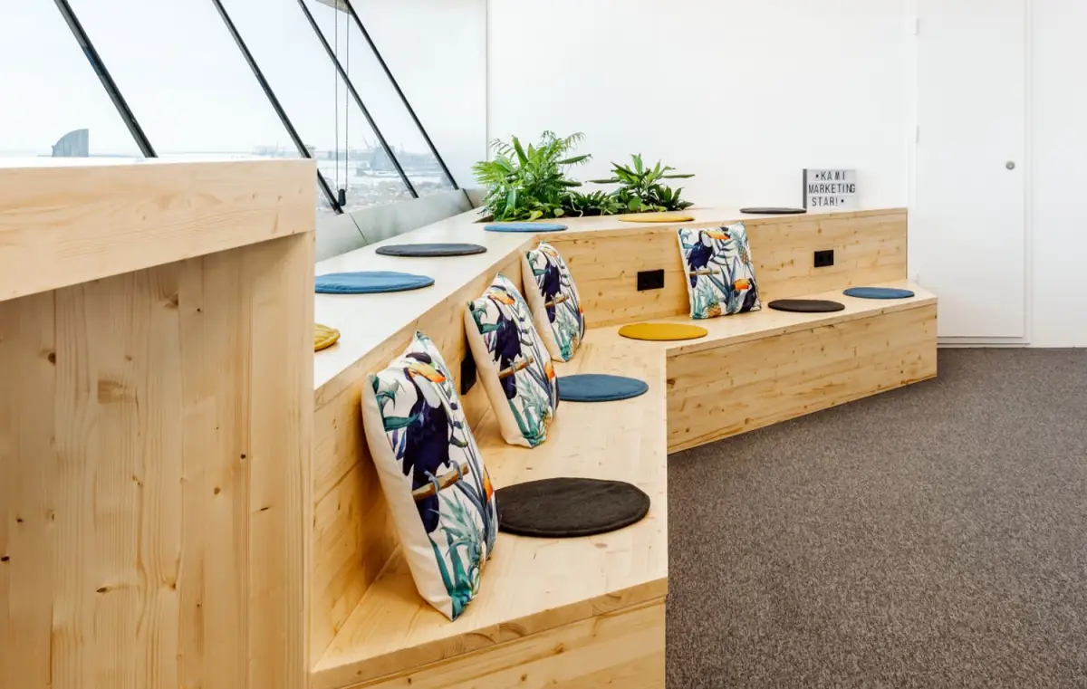 Pale timber built-in bench with patterned cushions and plants beneath a sloping window