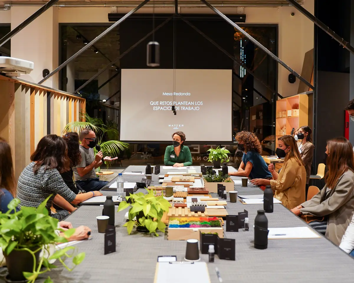 Speakers and attendees seated on both sides of a long table with material samples, facing a screen