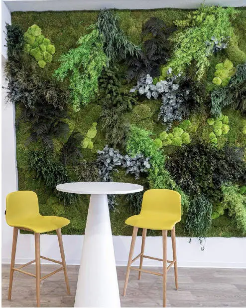 Vertical garden of preserved moss and plants beside a tall white table and yellow stools