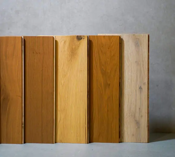 Cinco tablas de madera maciza en tonos que van del miel claro al marrón, apoyadas contra un muro de hormigón