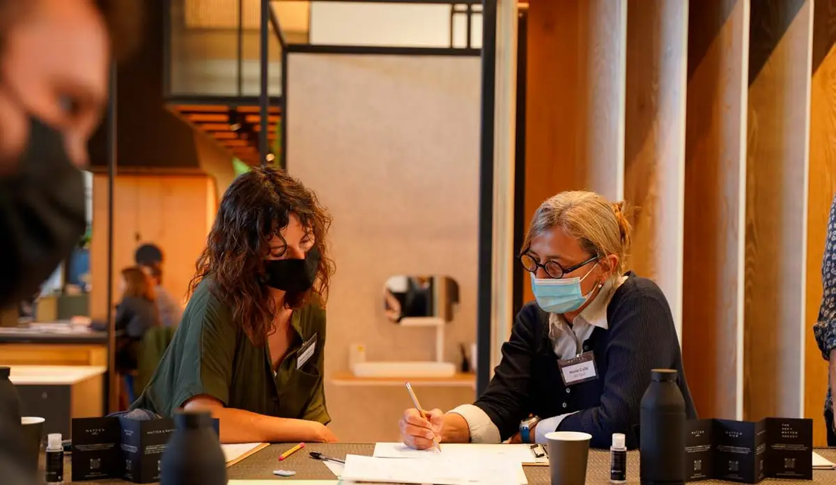 Dos participantes con mascarilla toman notas en la mesa de trabajo