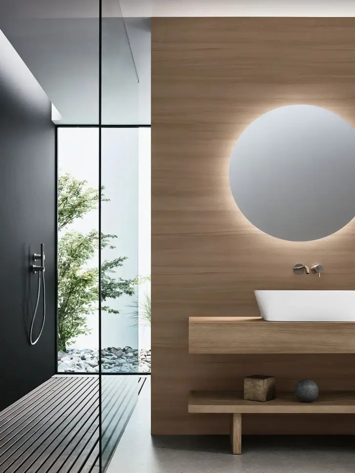 Bathroom with a white washbasin on a wooden countertop, a round backlit mirror and a shower opening onto a courtyard with a maple and pebbles.
