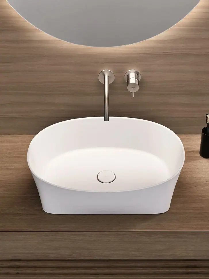 Top view of an oval white washbasin on a wooden countertop, with wall-mounted taps and a backlit mirror.