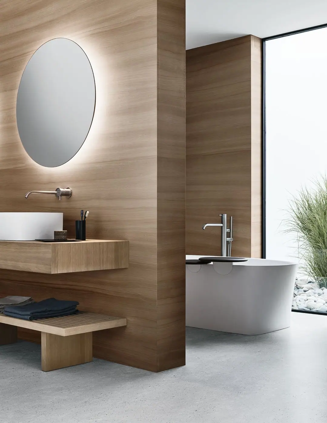 Bathroom with a white washbasin on a wooden countertop, an oval backlit mirror and, behind a wooden partition, a white freestanding bathtub with dark trays.