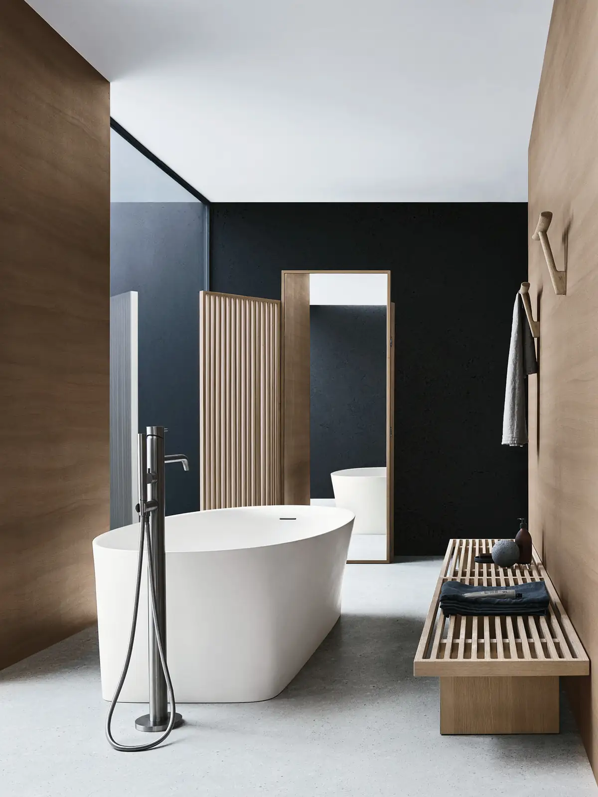 White freestanding bathtub between wooden panels, with a slatted and mirrored screen, a slatted bench and wooden coat racks on the wall.