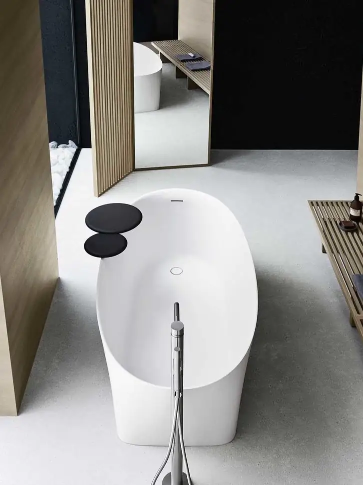 Overhead view of an oval white bathtub with two round black trays resting on the rim and a floor-standing tap.