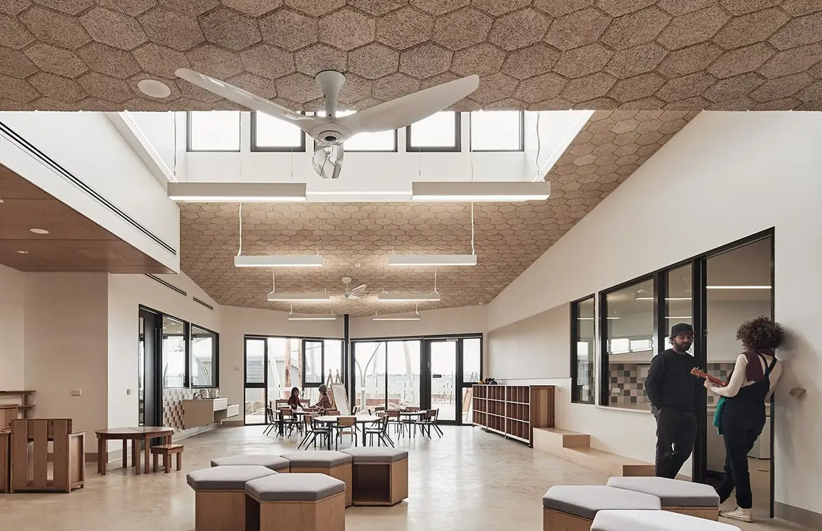 Espacio de educación infantil con techo de baldosas hexagonales, ventilador blanco, ventanas altas y asientos tapizados