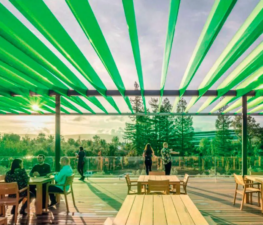 Terrace with wooden tables beneath a pergola of translucent green slats, with trees in the background