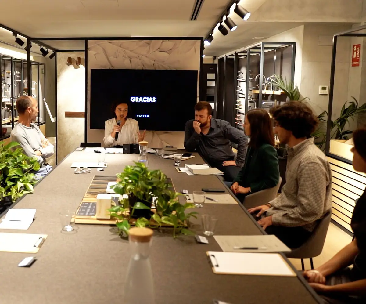 Speaker with a microphone at the head of a long table with materials and plants, surrounded by attendees