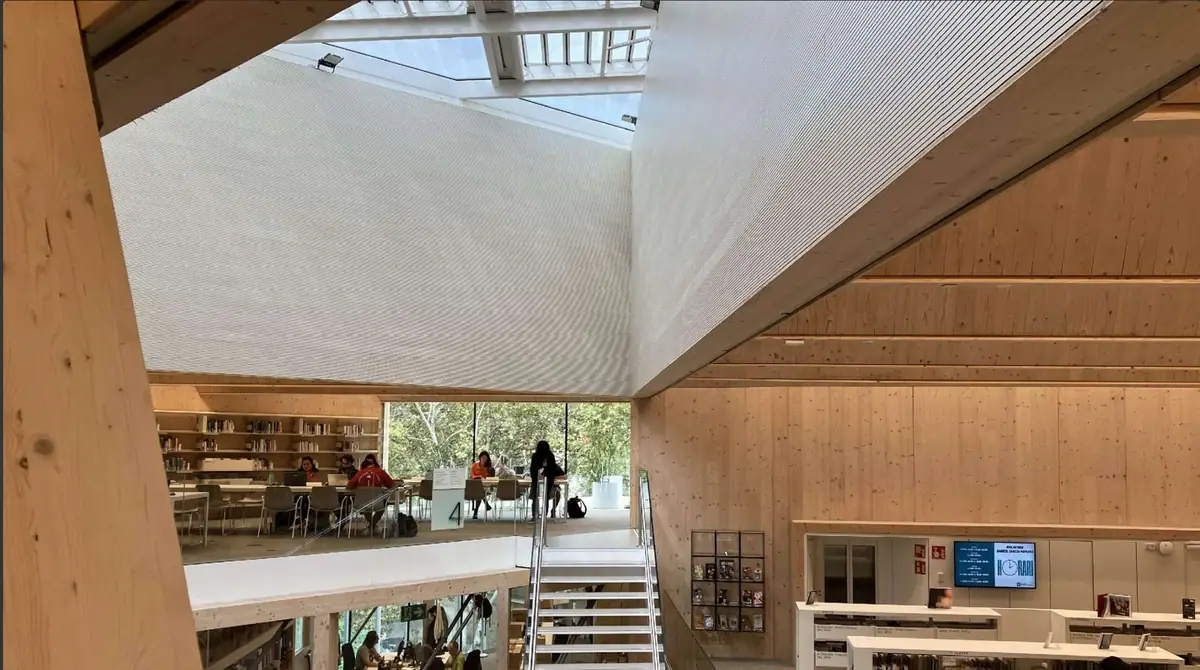 Atrio de una biblioteca con estructura de madera y paramentos superiores revestidos de lamas acústicas blancas bajo un lucernario