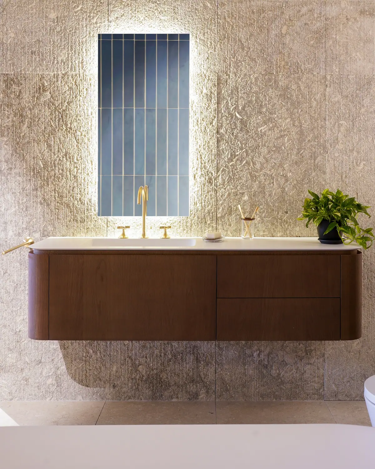 Mueble de baño suspendido de madera oscura con encimera blanca, grifería dorada y espejo retroiluminado sobre una pared de piedra texturizada