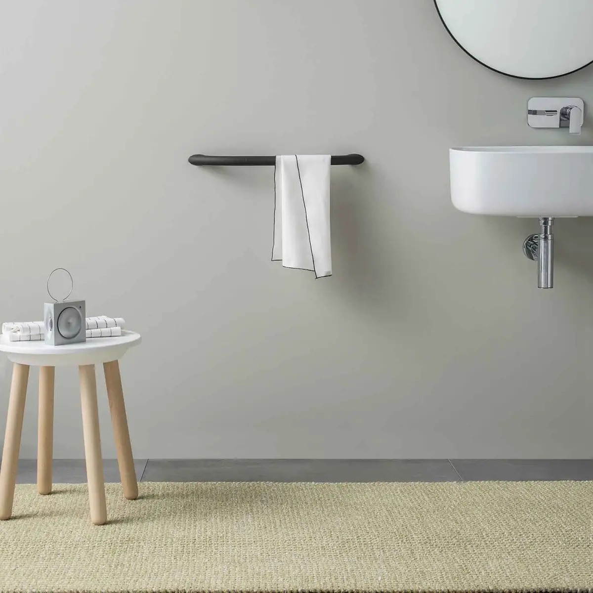 Toallero de seguridad gris antracita con una toalla blanca, junto a un lavabo mural, un espejo redondo y un taburete