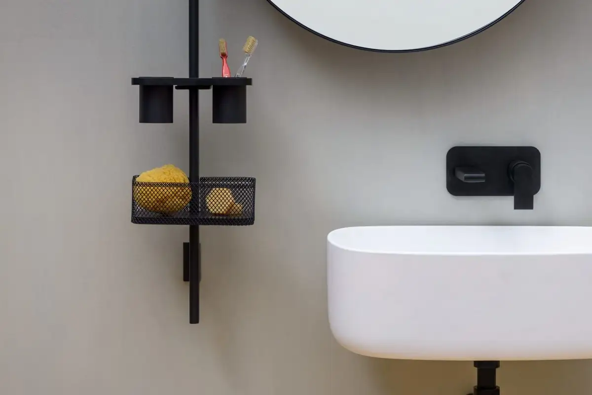 Barra vertical negra con vasos y cesta de rejilla junto a un lavabo blanco y un espejo redondo