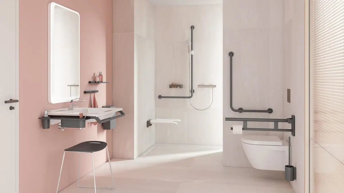 Baño accesible con paredes rosa, lavabo con barras, ducha con asiento abatible e inodoro con barra abatible en gris oscuro mate
