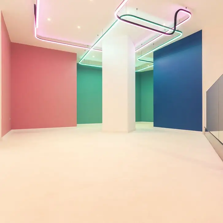 Sala con suelo continuo blanco brillante, paredes de color rosa, verde y azul y luminarias lineales de neón