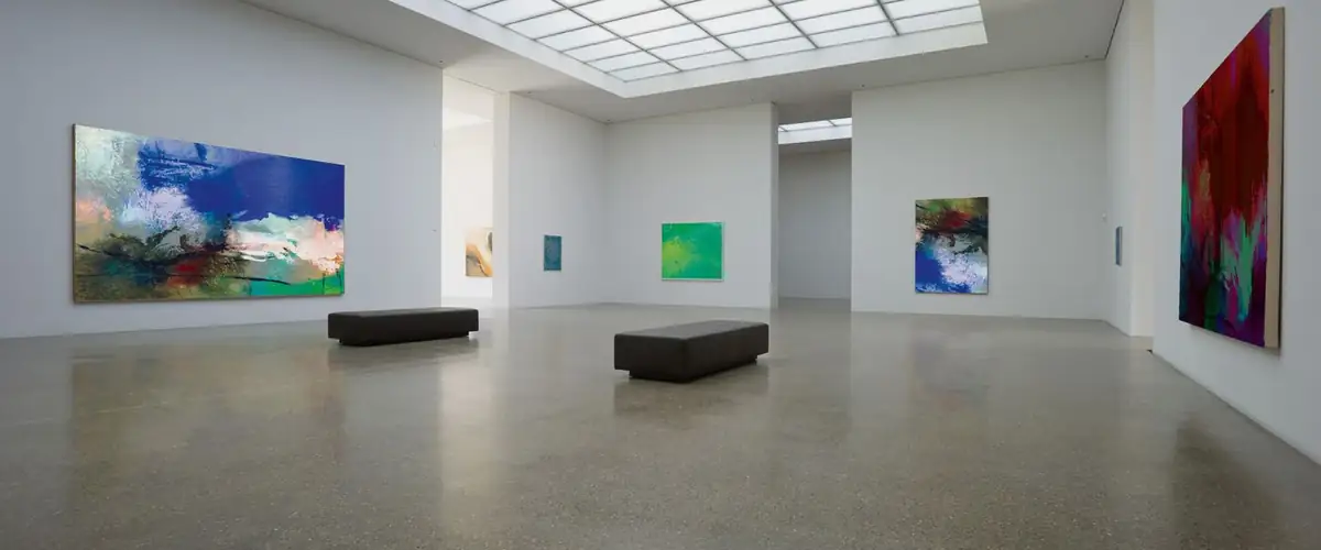 Sala de museo con pavimento continuo de terrazo gris claro, bancos oscuros y pinturas abstractas en las paredes