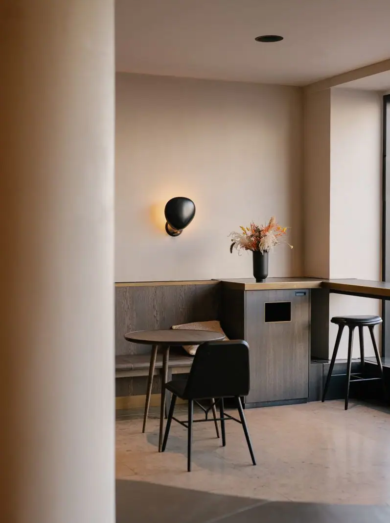 Zona de mesas de una cafetería con banco de madera oscura, aplique negro y paredes de yeso mineral claro