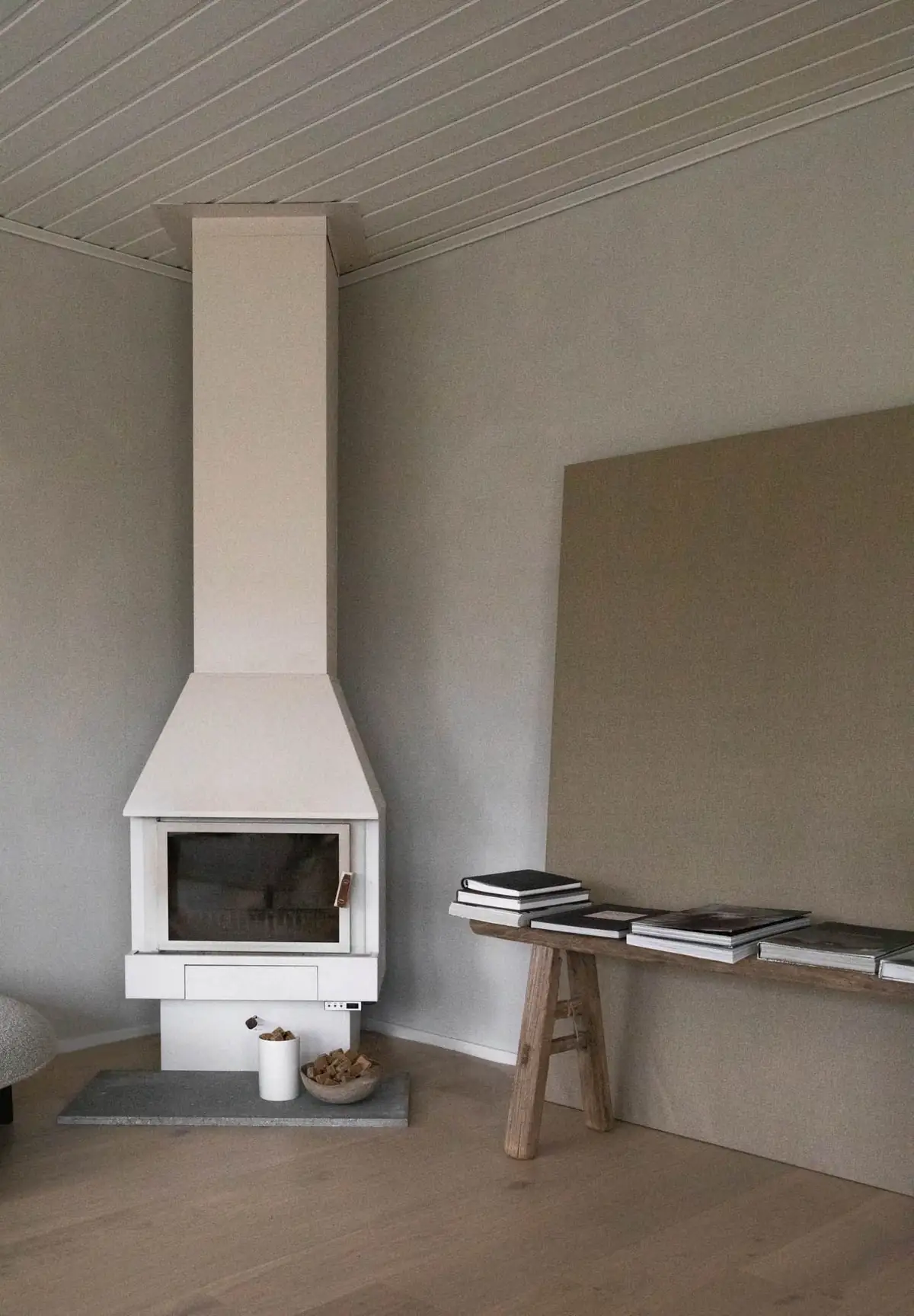 Sala con chimenea blanca en esquina, techo de lamas de madera, paredes de yeso gris y un gran lienzo apoyado en un banco