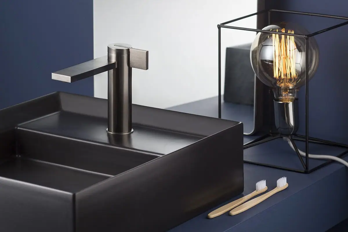 Mezclador monomando DOT316 de acabado oscuro sobre un lavabo negro rectangular, junto a una lámpara de filamento