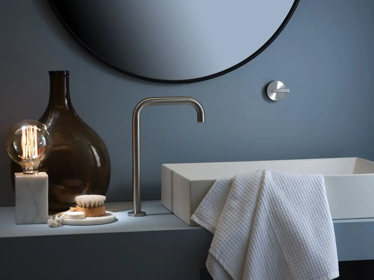 Caño de lavabo alto en acero cepillado junto a un mando rotativo empotrado en una pared gris azulada