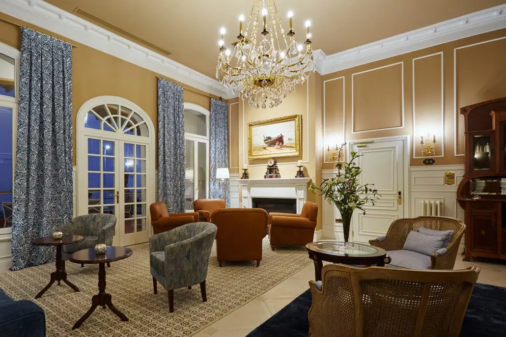 Salón de hotel con lámpara de araña, chimenea, sillones tapizados y molduras blancas en las paredes
