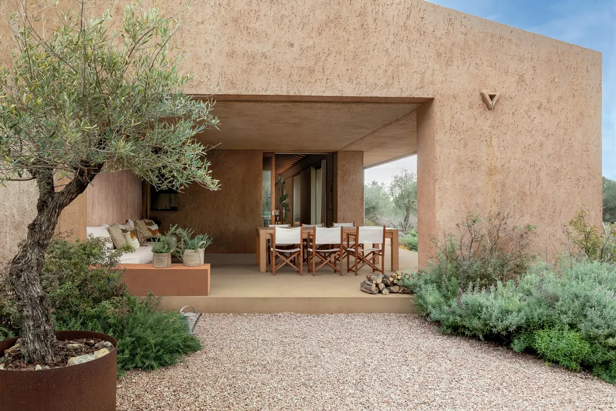 Porche exterior de una vivienda con paredes y pavimento de porcelánico de textura terrosa, mesa y olivo