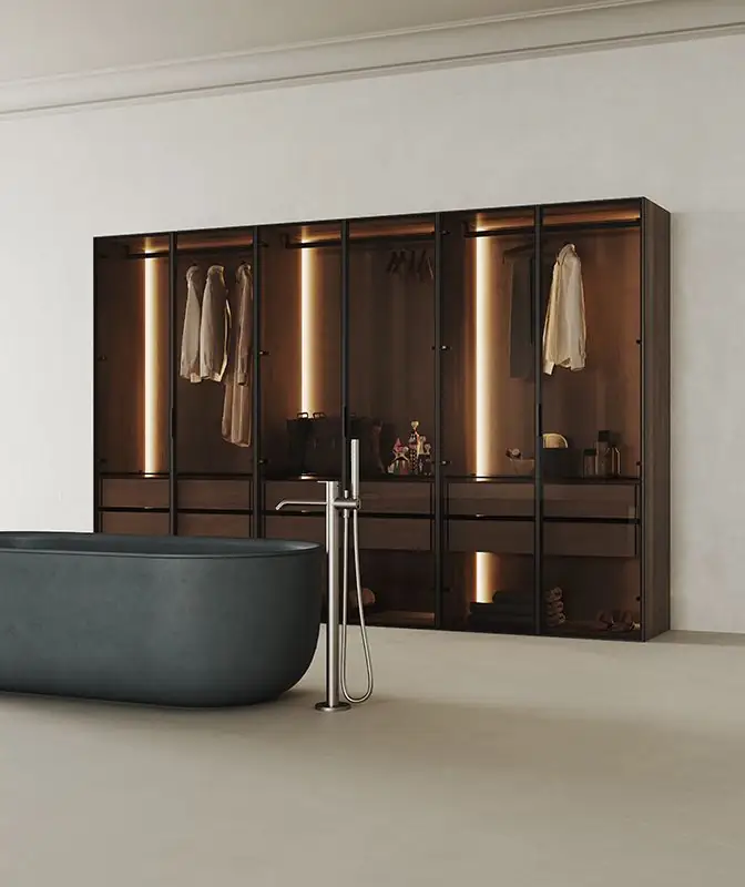 Walk-in wardrobe with glass doors and dark profiles, interior lighting and a grey freestanding bath in the foreground