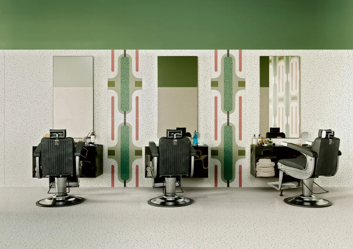 Barber’s shop with three chairs facing a speckled wall with decorated bands of green silhouettes and pink stripes between the mirrors.