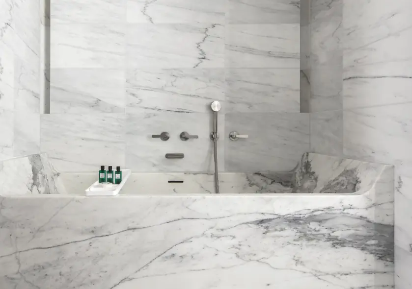 Bath clad in white veined marble with two handles, a cascade spout and a steel hand shower on the wall