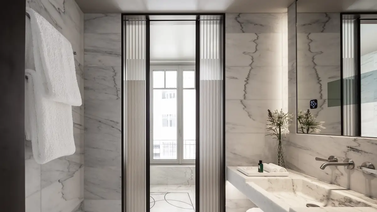 Bathroom clad in white veined marble with a marble basin and wall-mounted steel taps, next to a fluted glass door