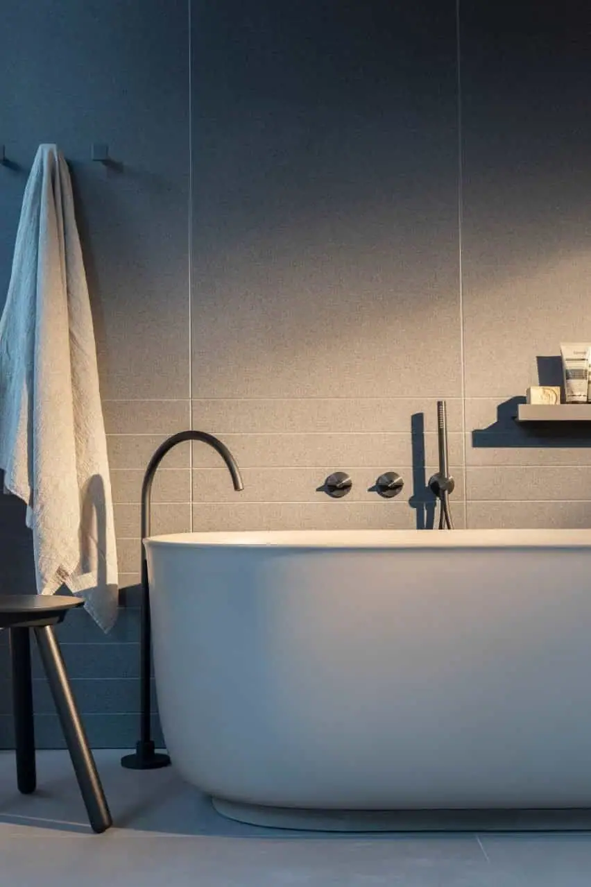White freestanding bath with a black floor-standing spout and wall-mounted controls and hand shower, next to a towel and a stool