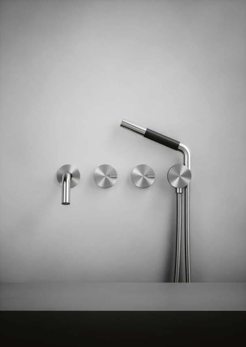 Wall-mounted spout, two circular controls and stainless steel hand shower on a grey wall