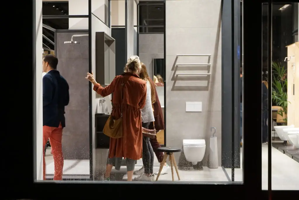 Guests looking at a bathroom setting with a wall-hung toilet and towel rail, seen through the glass