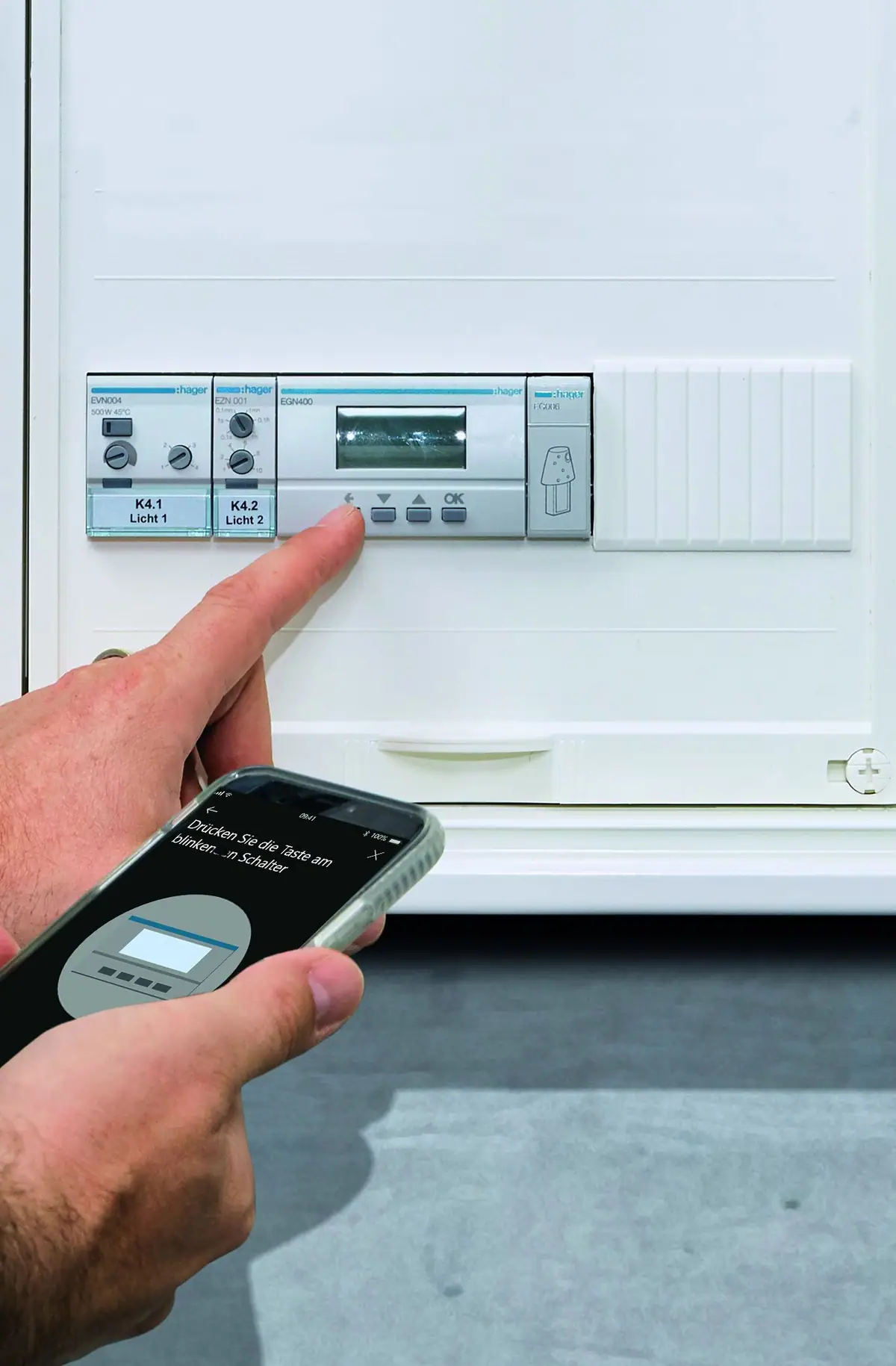 Hand pressing a Hager EGN400 digital time switch in a distribution board while the other holds a phone with the configuration app
