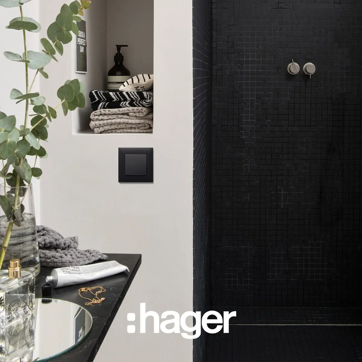 Black switch on a bathroom wall, beside a shower clad in black mosaic and a shelf with towels