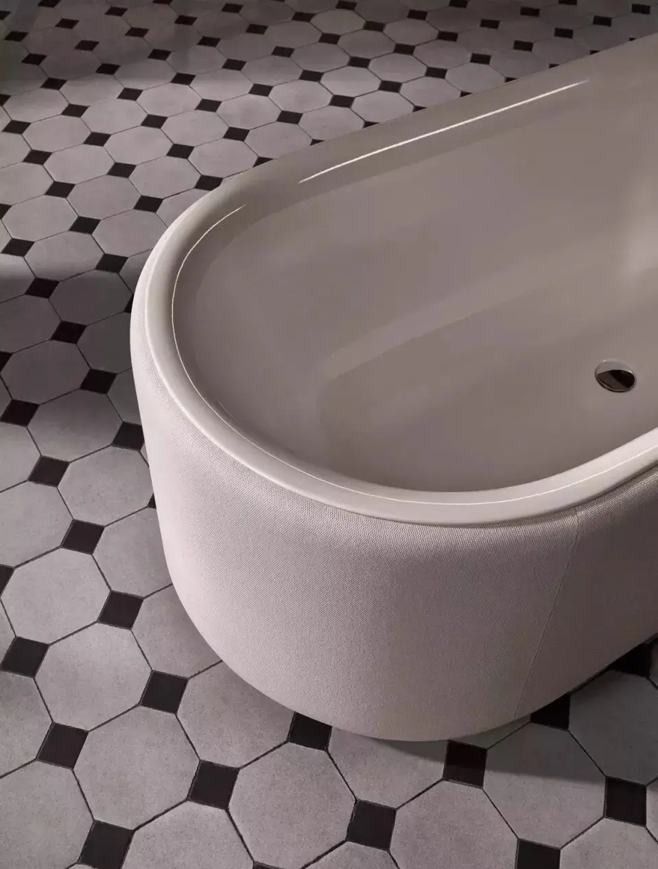 Detalle del borde de una bañera ovalada con interior esmaltado claro y faldón tapizado en tejido, sobre un suelo de baldosa octogonal