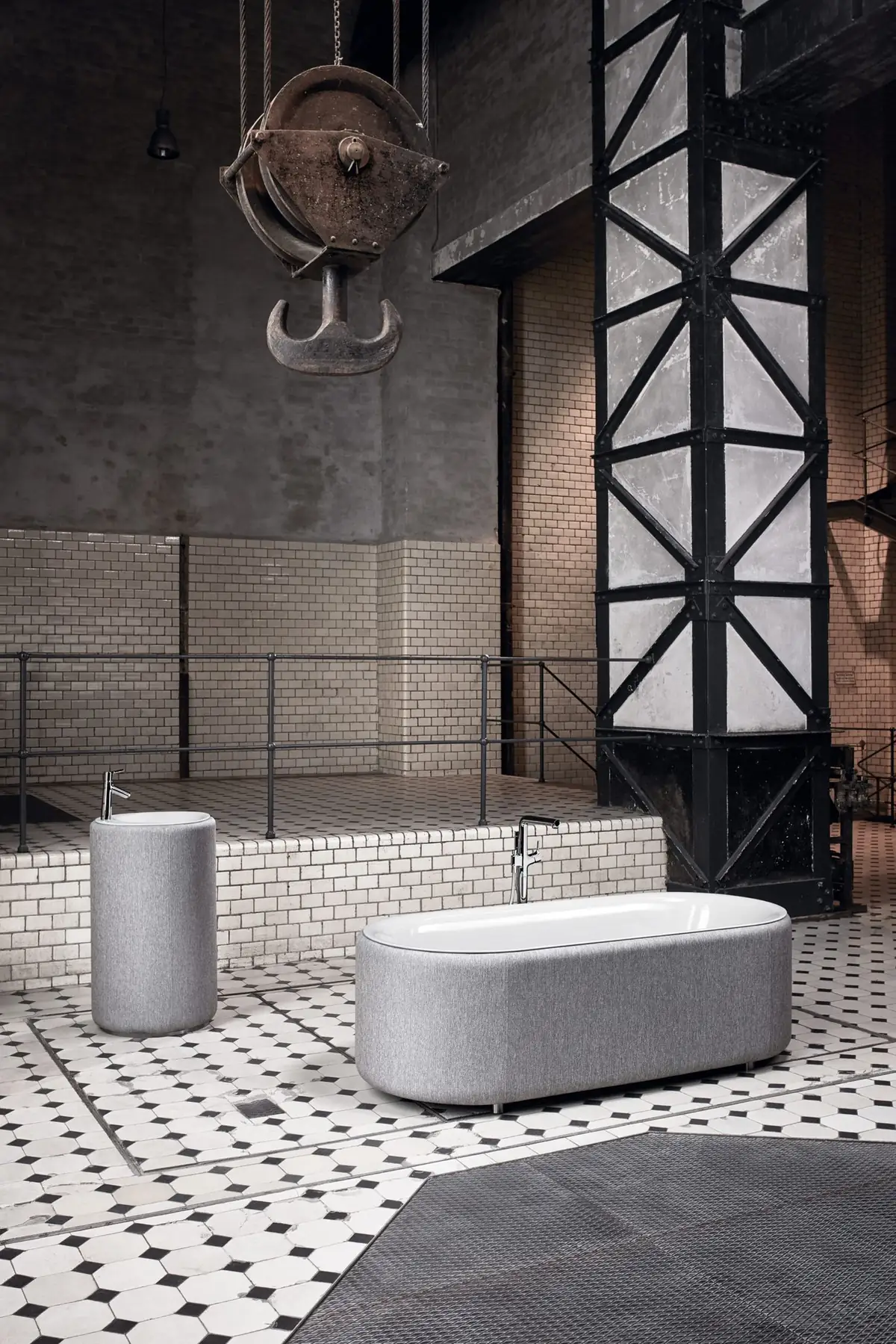Bañera ovalada y lavabo de pie tapizados en tejido gris claro, con grifería de pie cromada, bajo un gancho de grúa en una antigua sala de máquinas