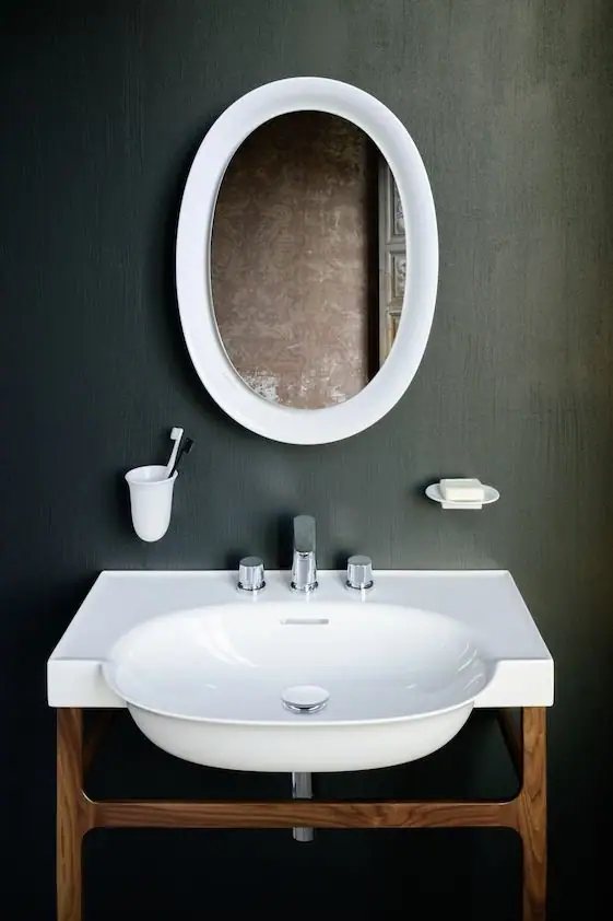 Lavabo blanco sobre bastidor de madera de nogal, con grifería de tres agujeros, espejo ovalado de marco blanco y accesorios cerámicos en una pared oscura