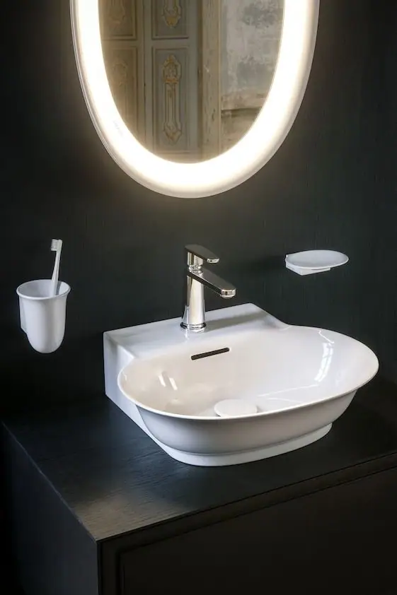 Lavabo blanco de formas curvas con grifo monomando sobre un mueble oscuro, bajo un espejo ovalado iluminado
