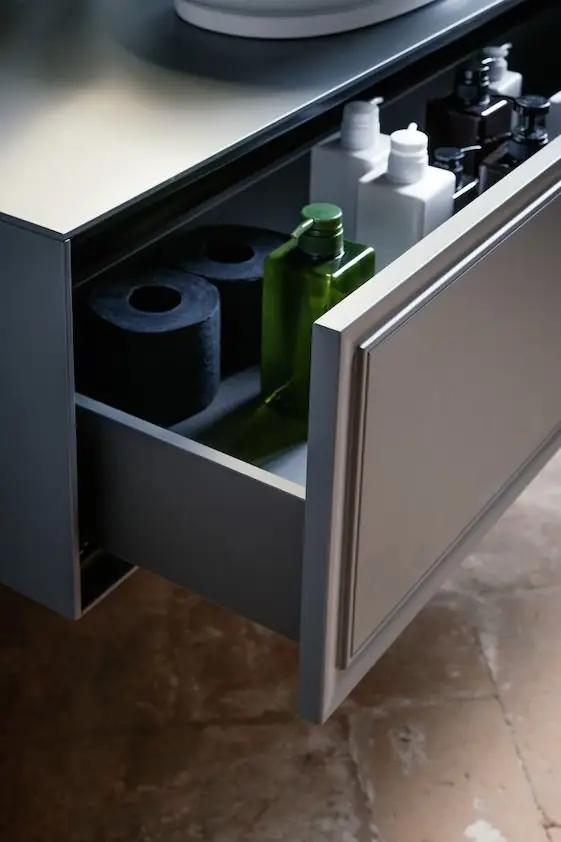 Detalle de un cajón abierto de un mueble de baño gris oscuro con frente moldurado y productos de aseo en su interior