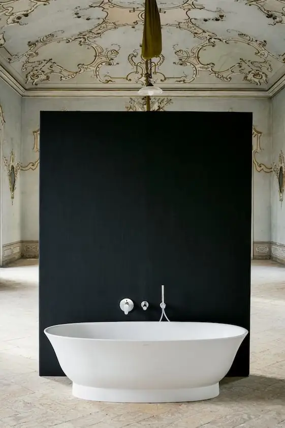 Bañera exenta blanca de bordes finos ante un panel negro, con grifería mural cromada, en una sala con techo ornamentado