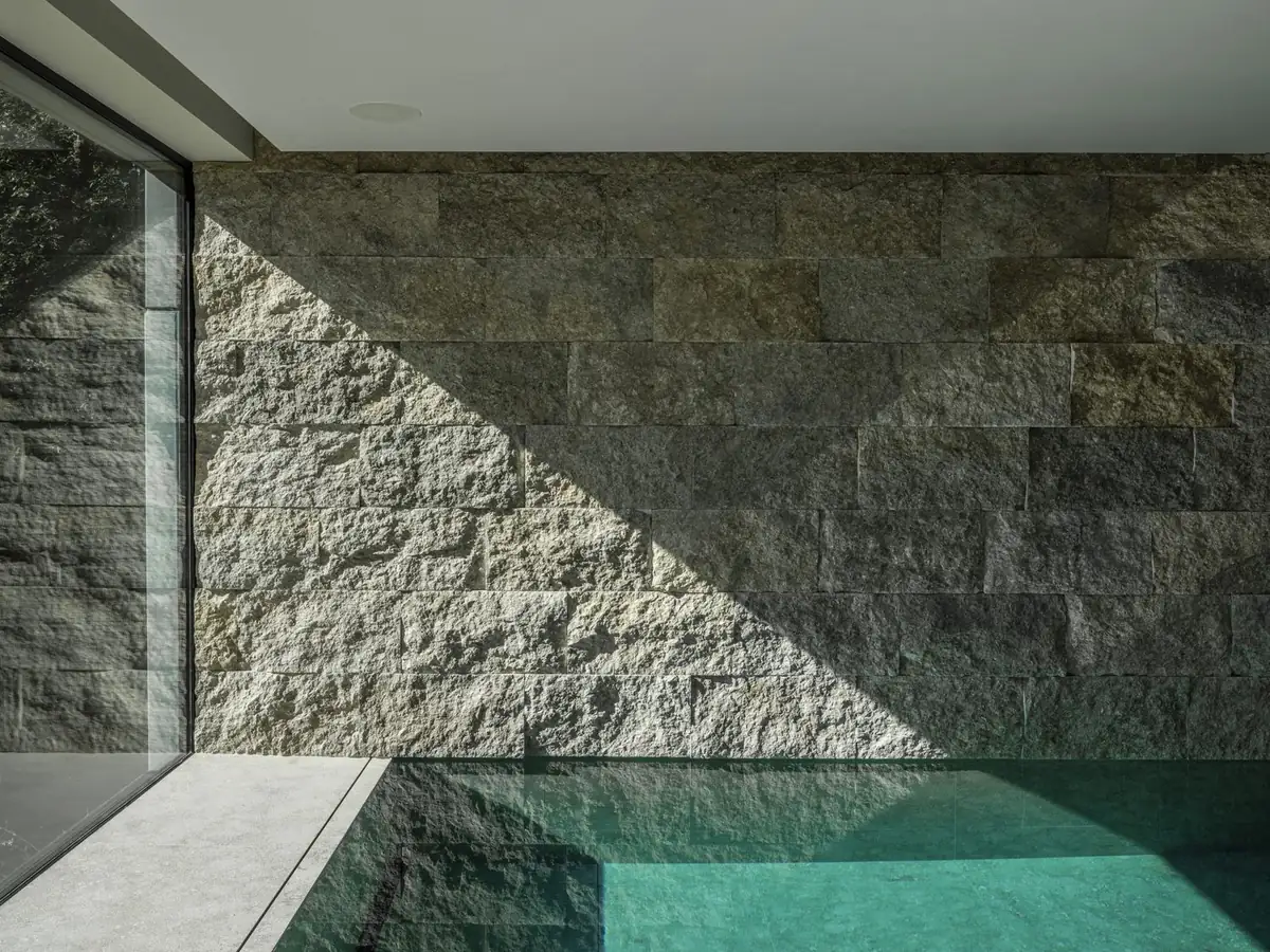 Muro de piedra gris en acabado partido junto a una piscina cubierta, con una diagonal de luz y sombra