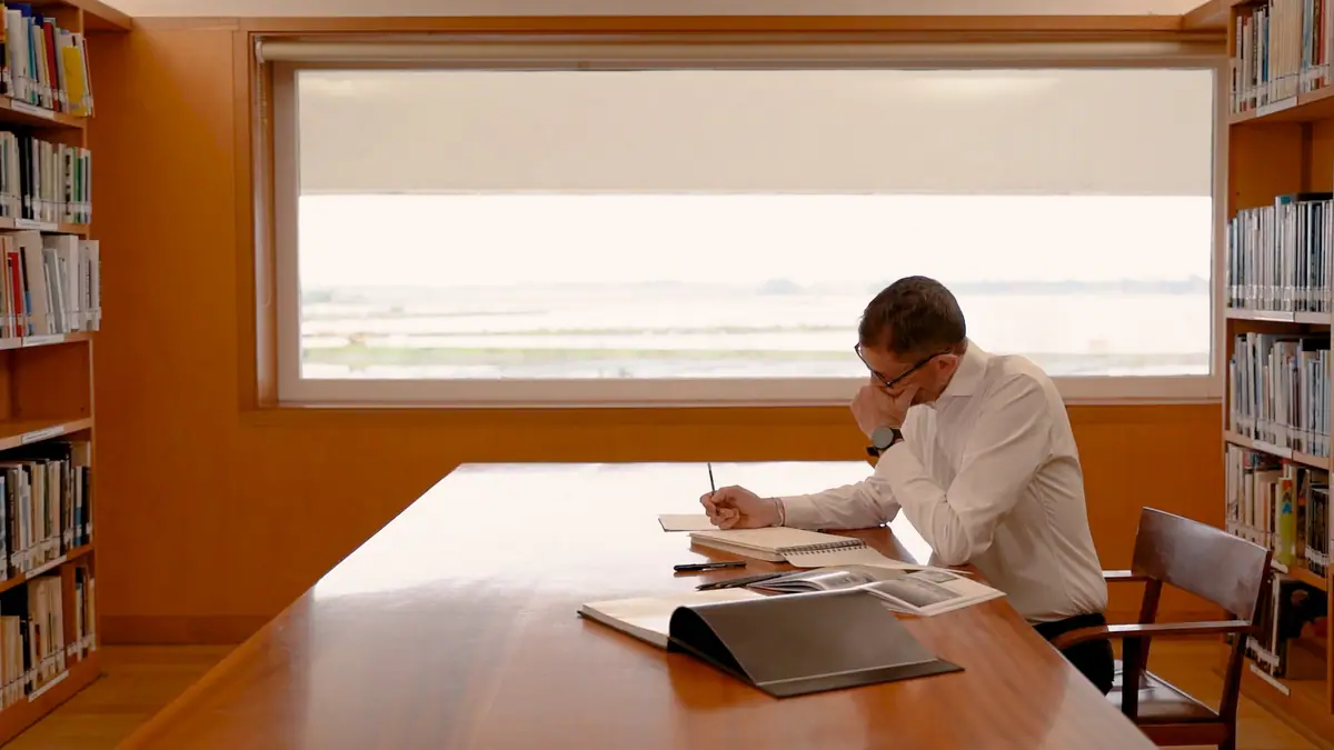 Man in a white shirt drawing at a wooden table in a library, in front of a large window