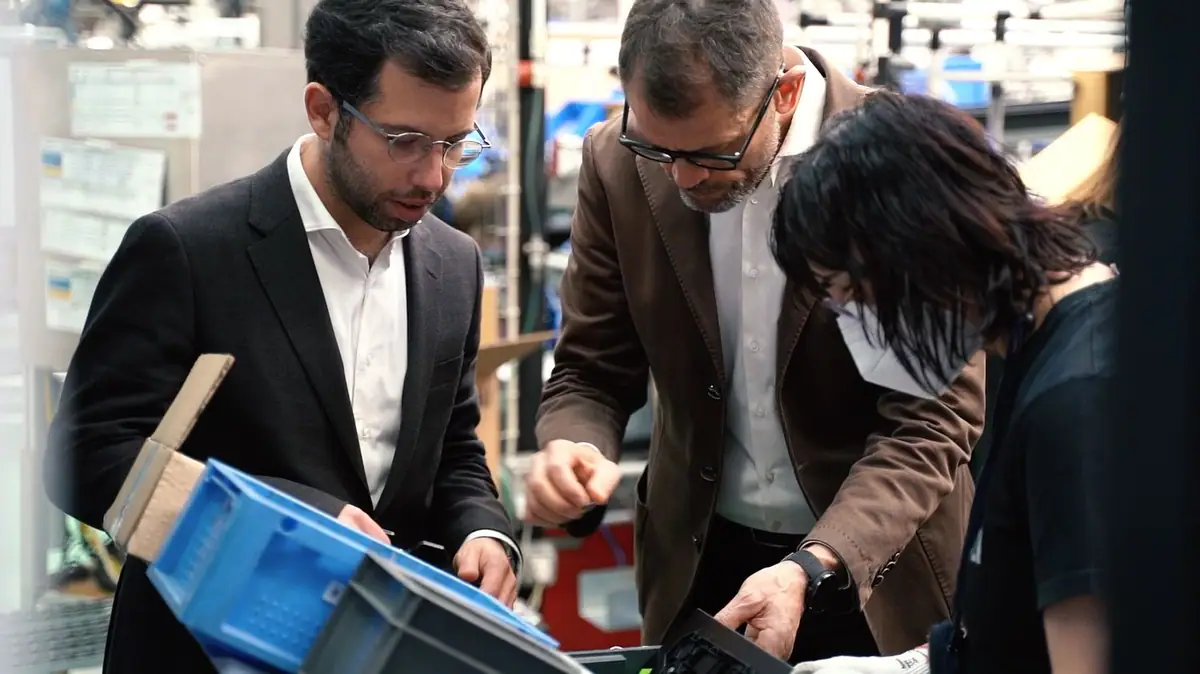 Two men and a female worker check components in plastic crates in an industrial plant