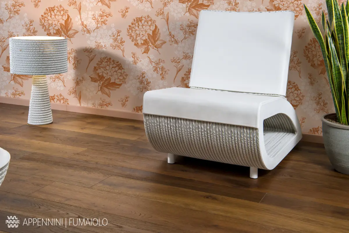 White armchair and cardboard lamp on a floor of warm brown oak boards, against a floral wallpaper