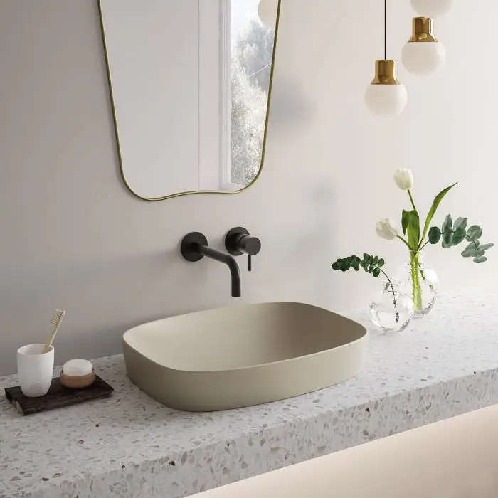 Softly contoured ceramic washbasin on a terrazzo worktop, with black wall-mounted taps and a gold-framed mirror