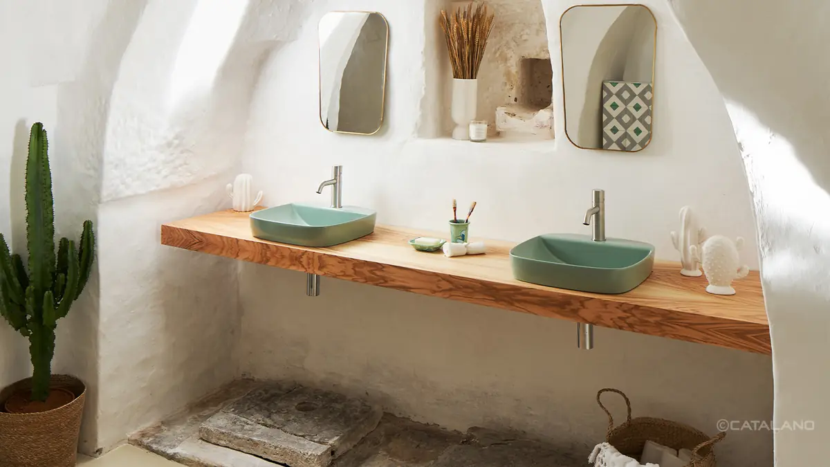Two green thin-edged washbasins on a wooden worktop, beneath two gold-framed mirrors on a whitewashed wall