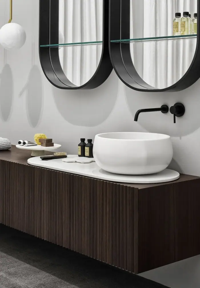 Lavabo de apoyo redondo blanco sobre un top ovalado blanco y un mueble suspendido de madera oscura con lamas, bajo dos espejos ovalados