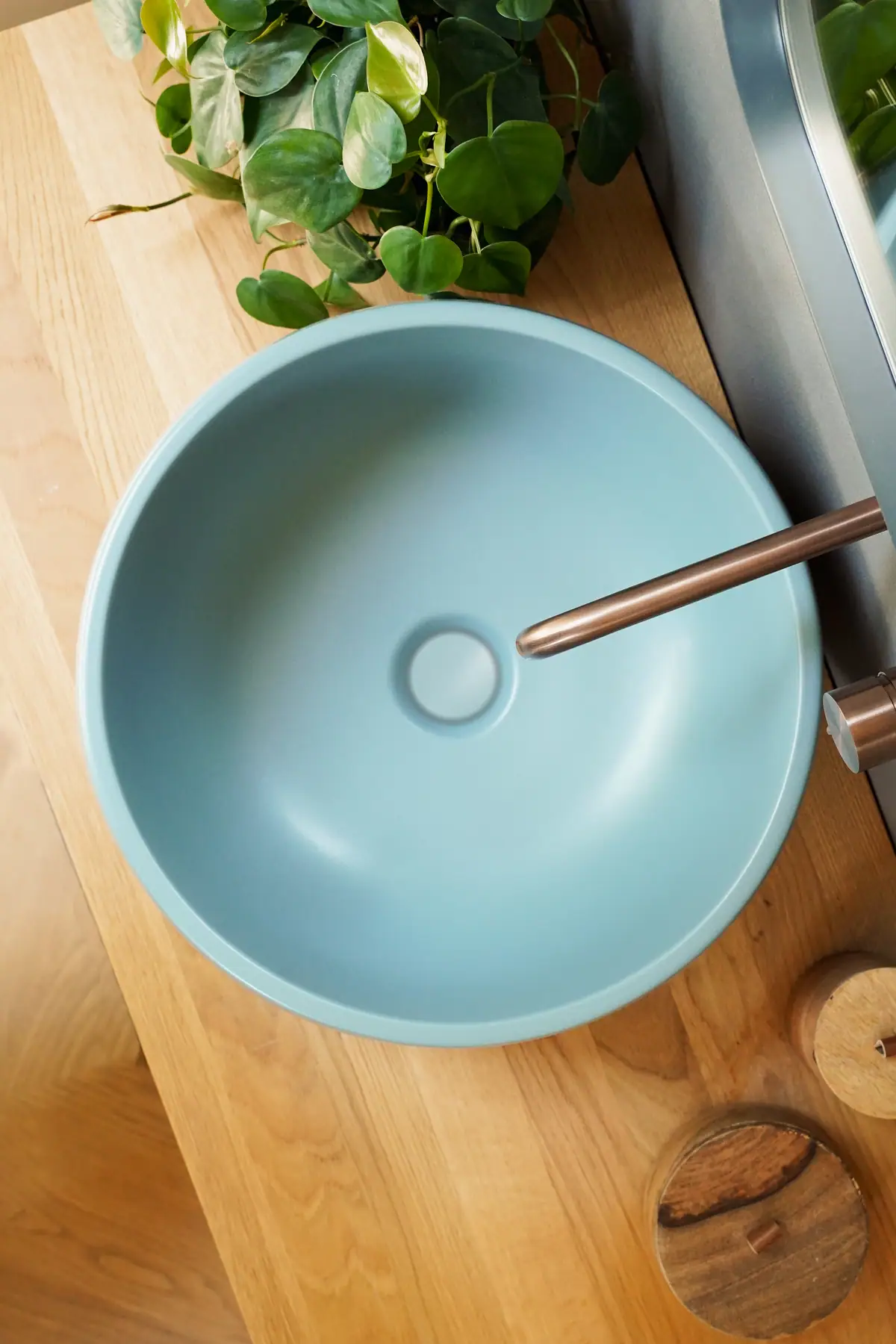 Vista cenital de un lavabo de apoyo redondo azul sobre una encimera de madera, con un caño de bronce y una planta